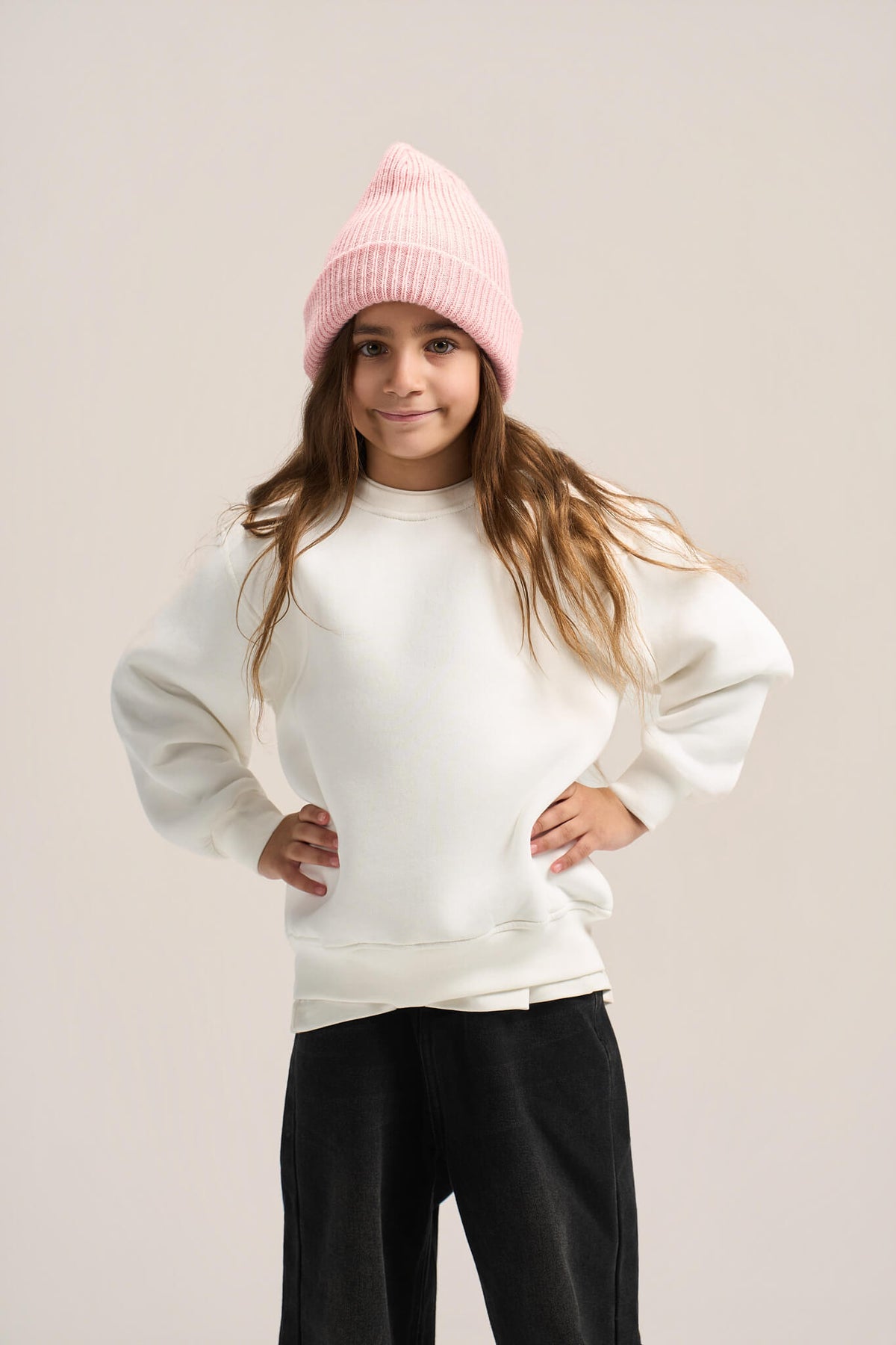A young girl wearing a light pink ribbed knit beanie, a white oversized crewneck sweatshirt, and black sweatpants, posing against a solid light beige studio background.