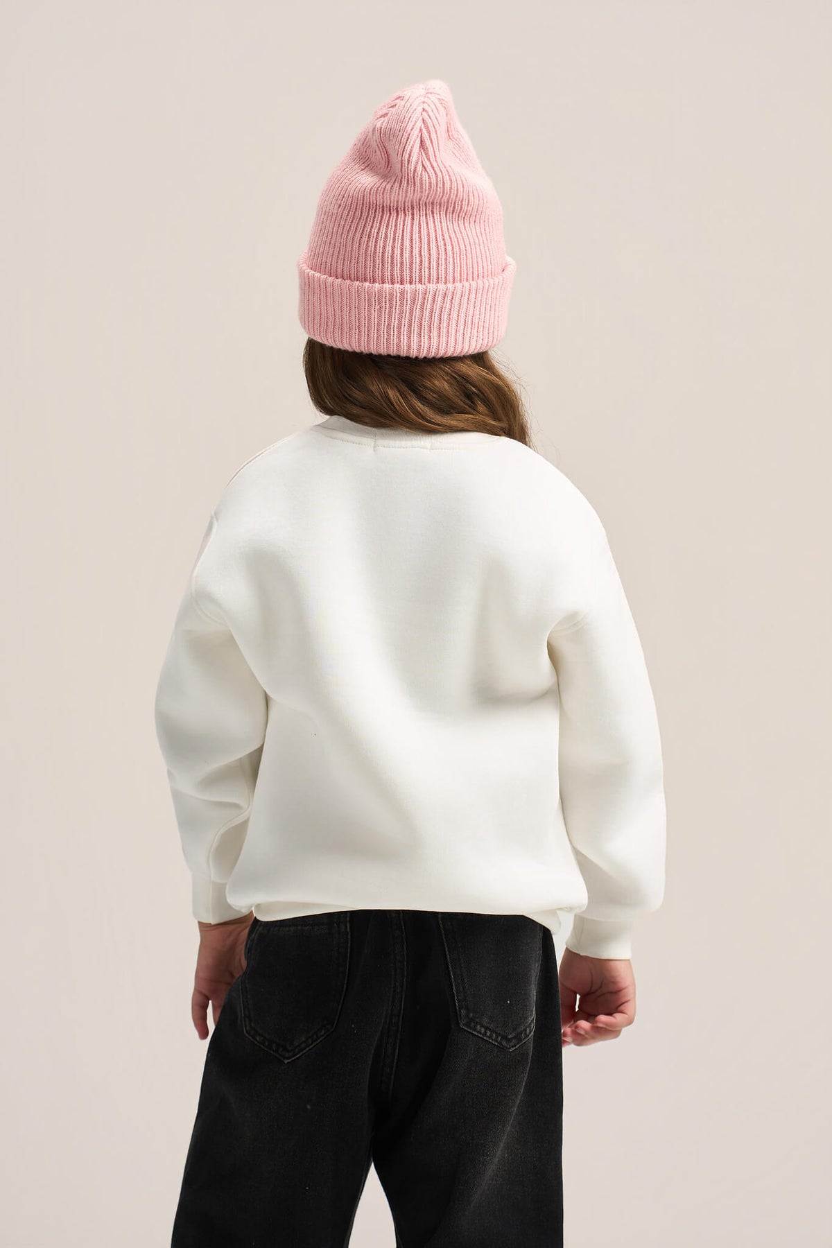 A child shown from the back wearing a cream-colored crewneck sweatshirt, black pants, and a pink ribbed knit beanie against a solid light beige background.
