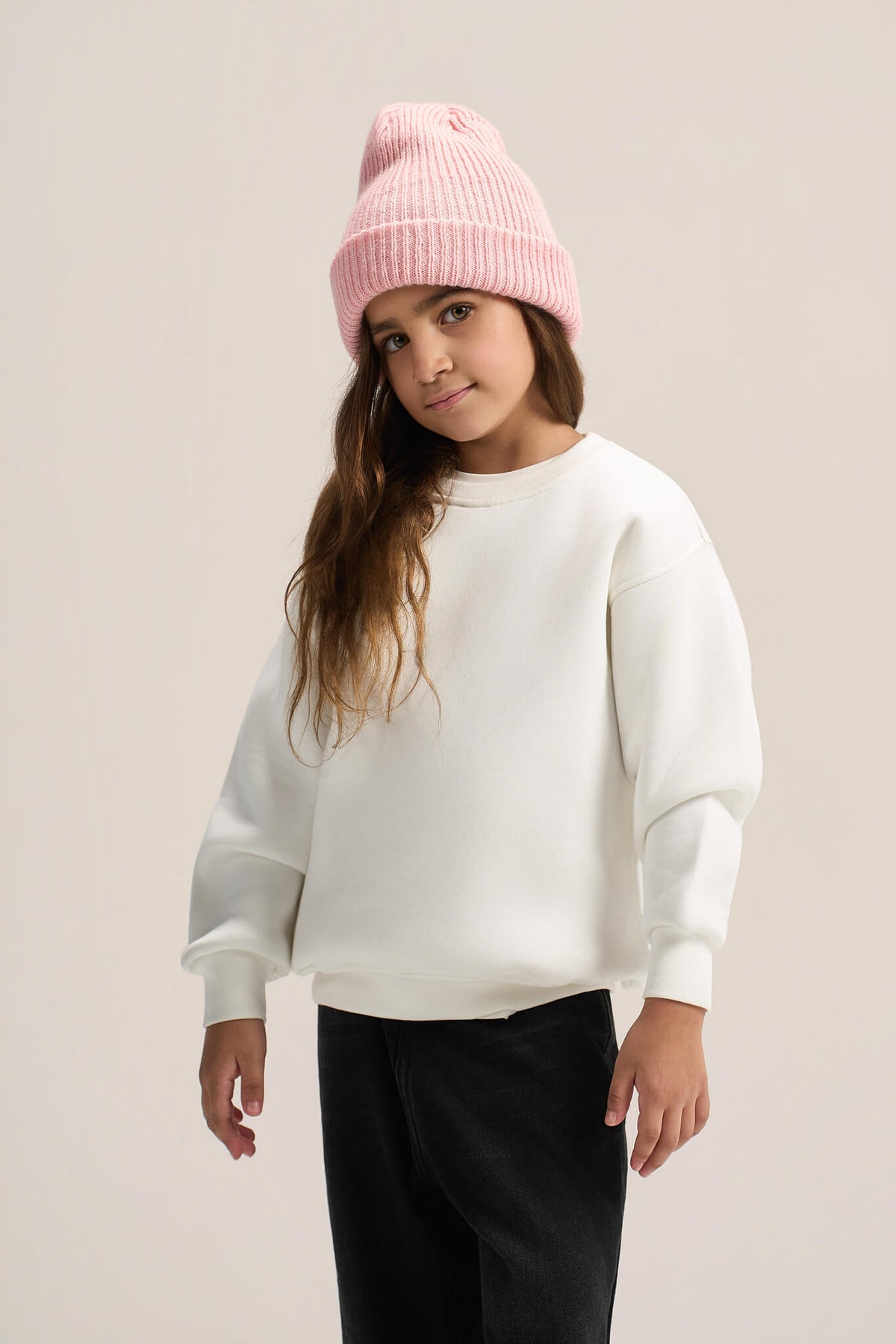 A young girl wearing a light pink ribbed knit beanie, a plain white crewneck sweatshirt, and black pants, standing against a solid light beige studio background.