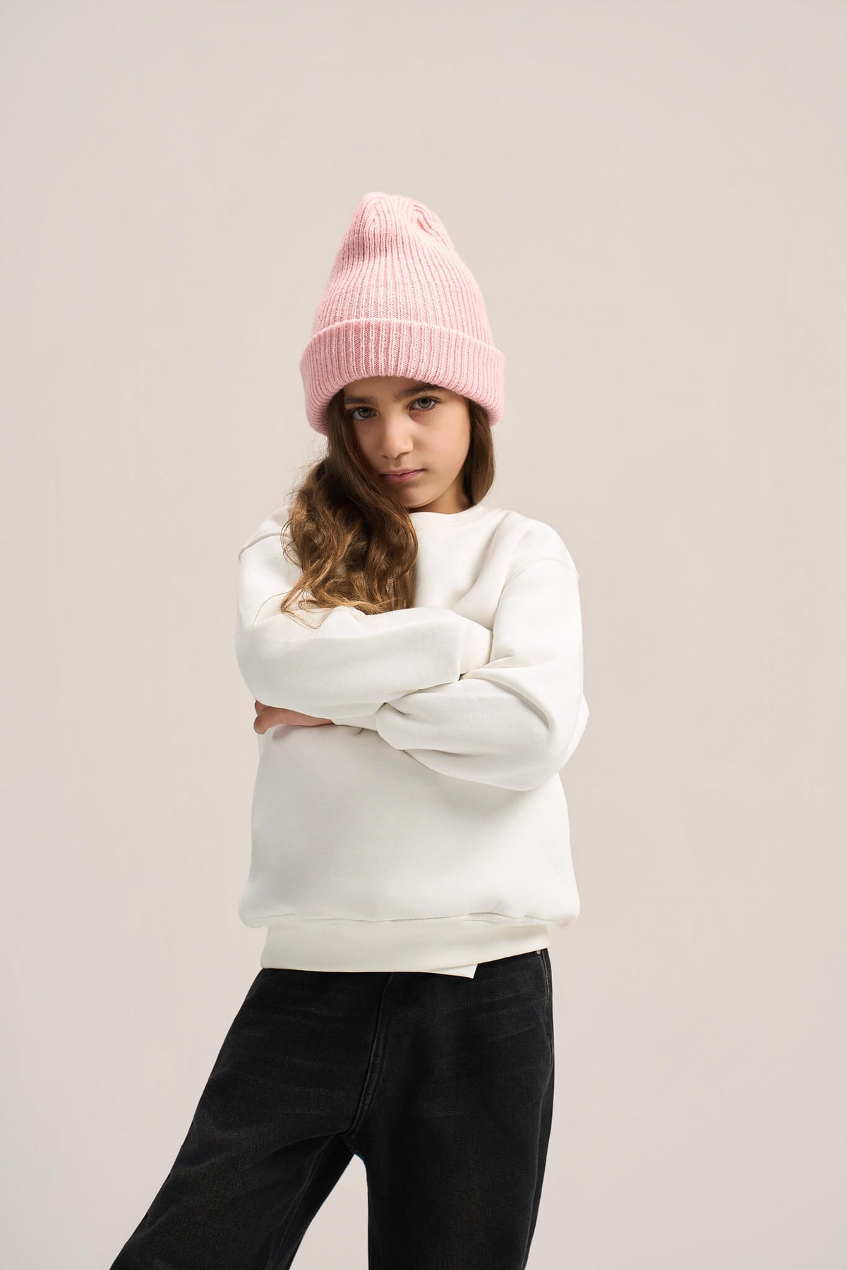 A child model wearing a light pink ribbed knit beanie, a white crewneck sweatshirt, and black pants, posed against a solid light beige studio background.