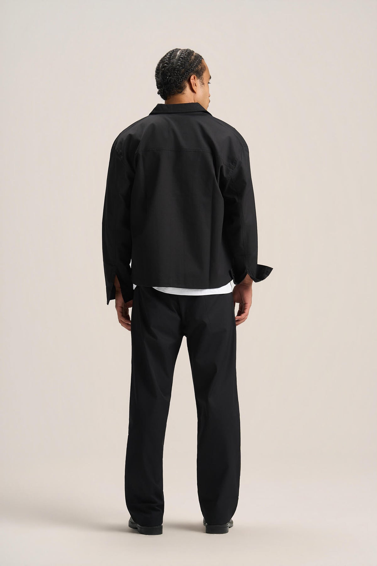 A man viewed from the back wearing a black long-sleeve button-up shirt, black trousers, and black shoes, standing against a plain light beige studio background.