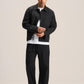 A man modeling a black jacket and matching black trousers over a white t-shirt, standing against a solid light beige studio background.