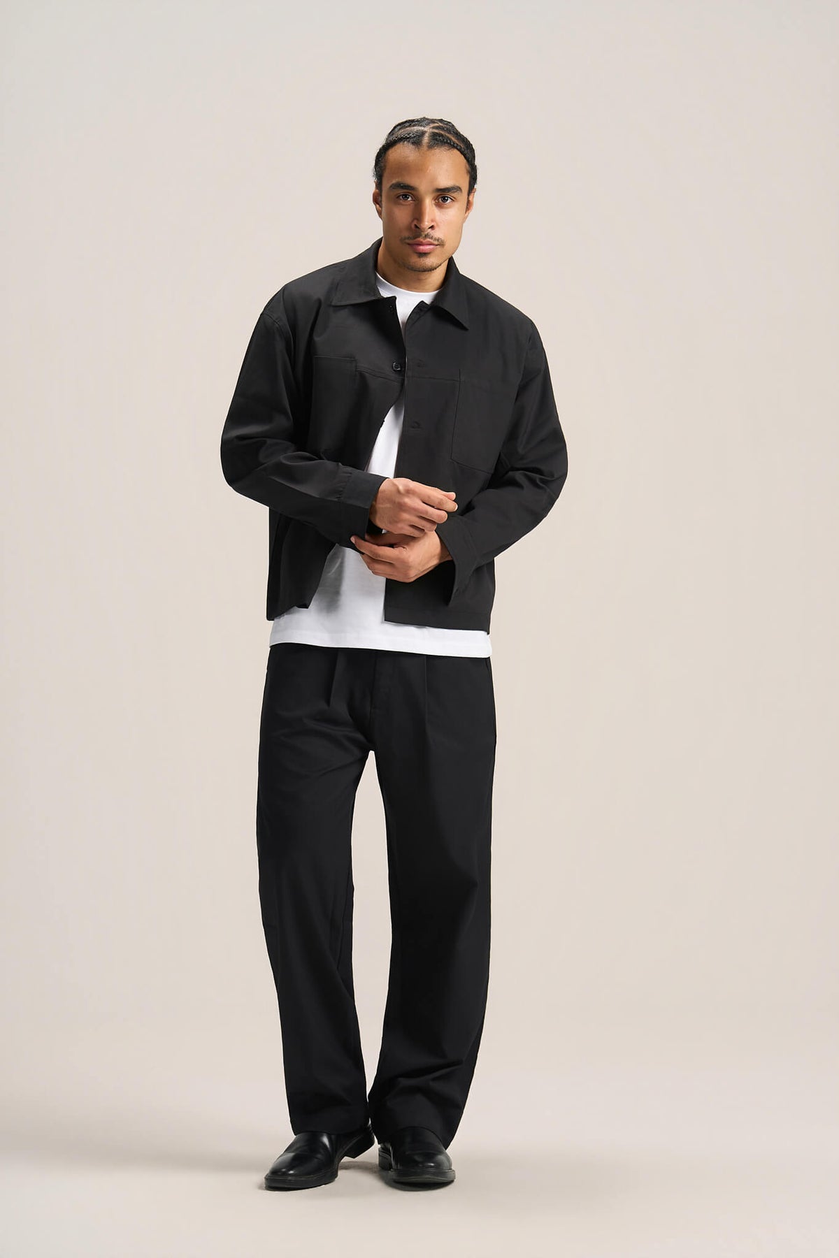 A man modeling a black jacket and matching black trousers over a white t-shirt, standing against a solid light beige studio background.