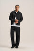 A man modeling a black jacket and matching black trousers over a white t-shirt, standing against a solid light beige studio background.