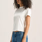 A woman with curly dark hair wearing a white short-sleeve crop top and blue jeans, standing against a plain light beige studio background.