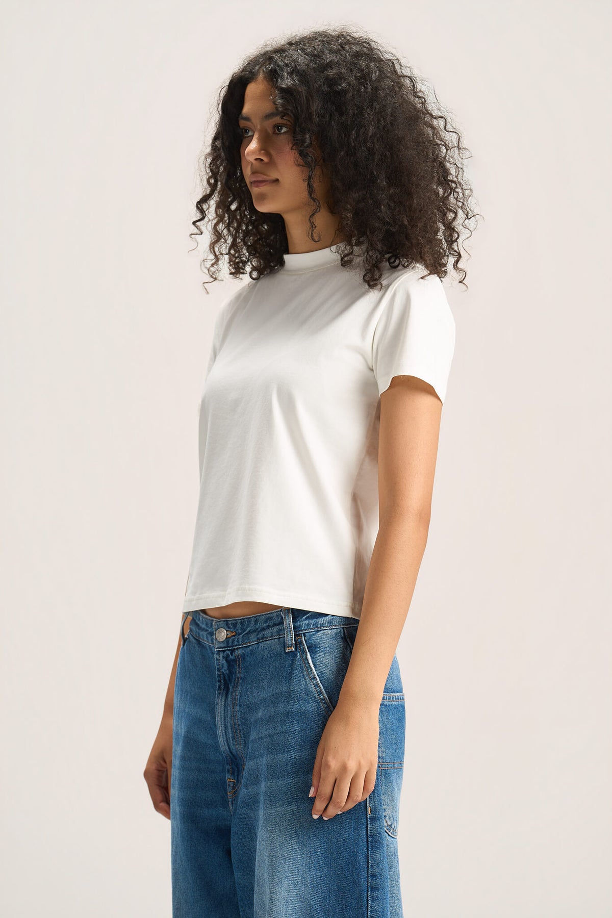 A woman with curly dark hair wearing a white short-sleeve crop top and blue jeans, standing against a plain light beige studio background.