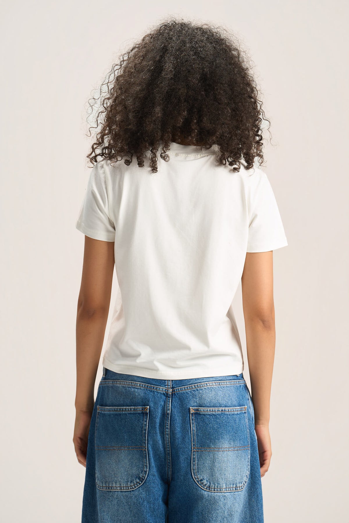 A person shown from the back wearing a plain white short-sleeve T-shirt and blue jeans against a light neutral studio background.
