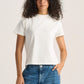 A woman with curly dark hair wearing a plain white short-sleeve crewneck t-shirt and blue jeans, standing against a light beige studio background.