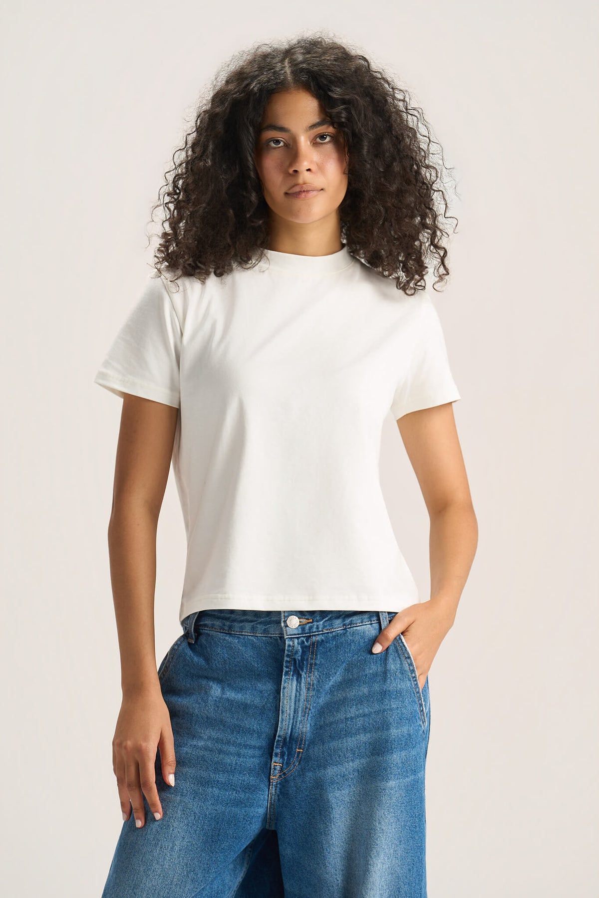 A woman with curly dark hair wearing a plain white short-sleeve crewneck t-shirt and blue jeans, standing against a light beige studio background.
