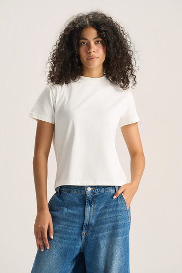 A woman with curly dark hair wearing a plain white short-sleeve crewneck t-shirt and blue jeans, standing against a light beige studio background.