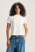 A woman with curly dark hair wearing a plain white short-sleeve crewneck t-shirt and blue jeans, standing against a light beige studio background.