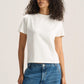 A woman with curly dark hair wearing a plain white short-sleeve crewneck t-shirt and blue jeans, standing against a light beige studio background.