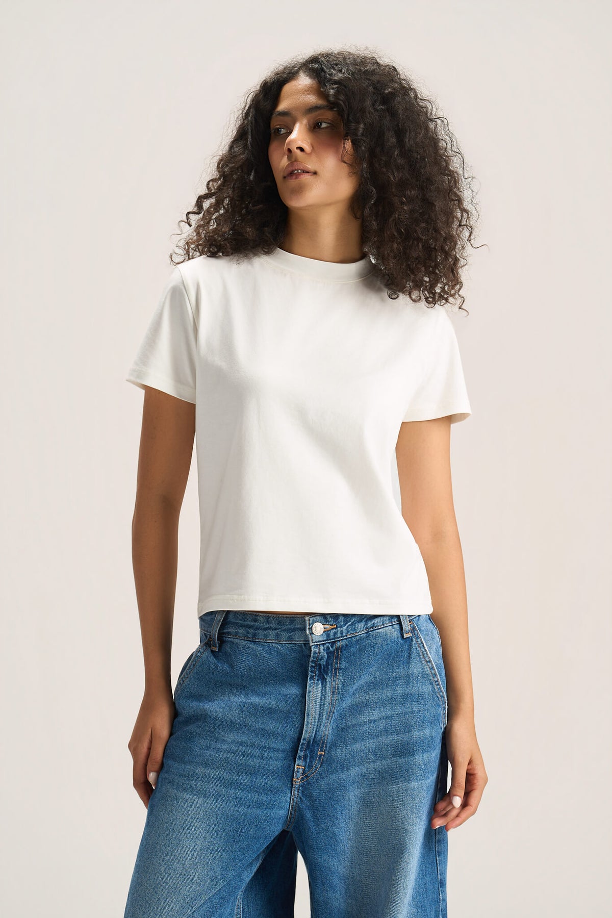 A woman with curly dark hair wearing a plain white short-sleeve crewneck t-shirt and blue jeans, standing against a light beige studio background.