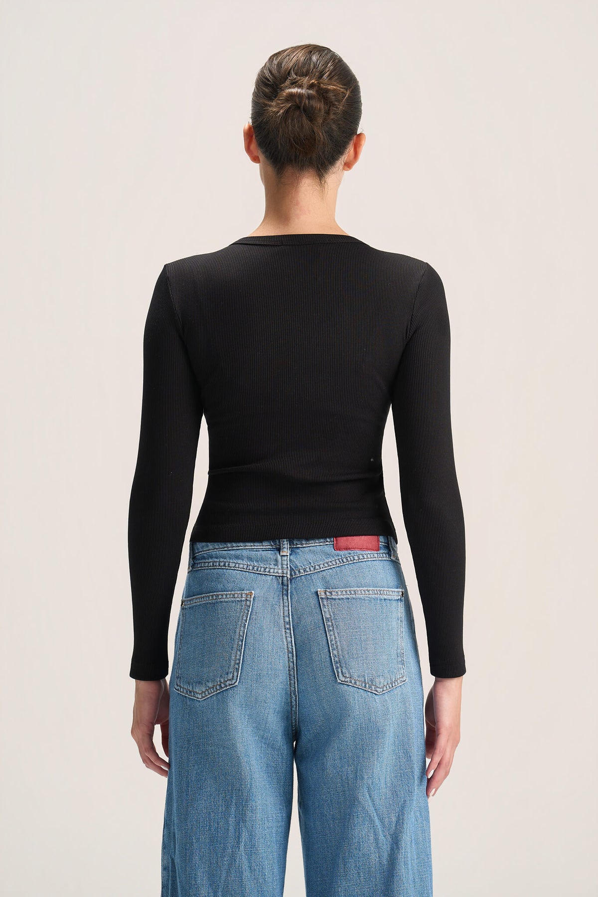A woman shown from the back wearing a fitted black long-sleeve top and light blue jeans against a plain light background.