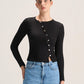 A model wearing a black long-sleeve knit top with a decorative button-down front and light blue jeans, photographed against a plain light gray studio background.