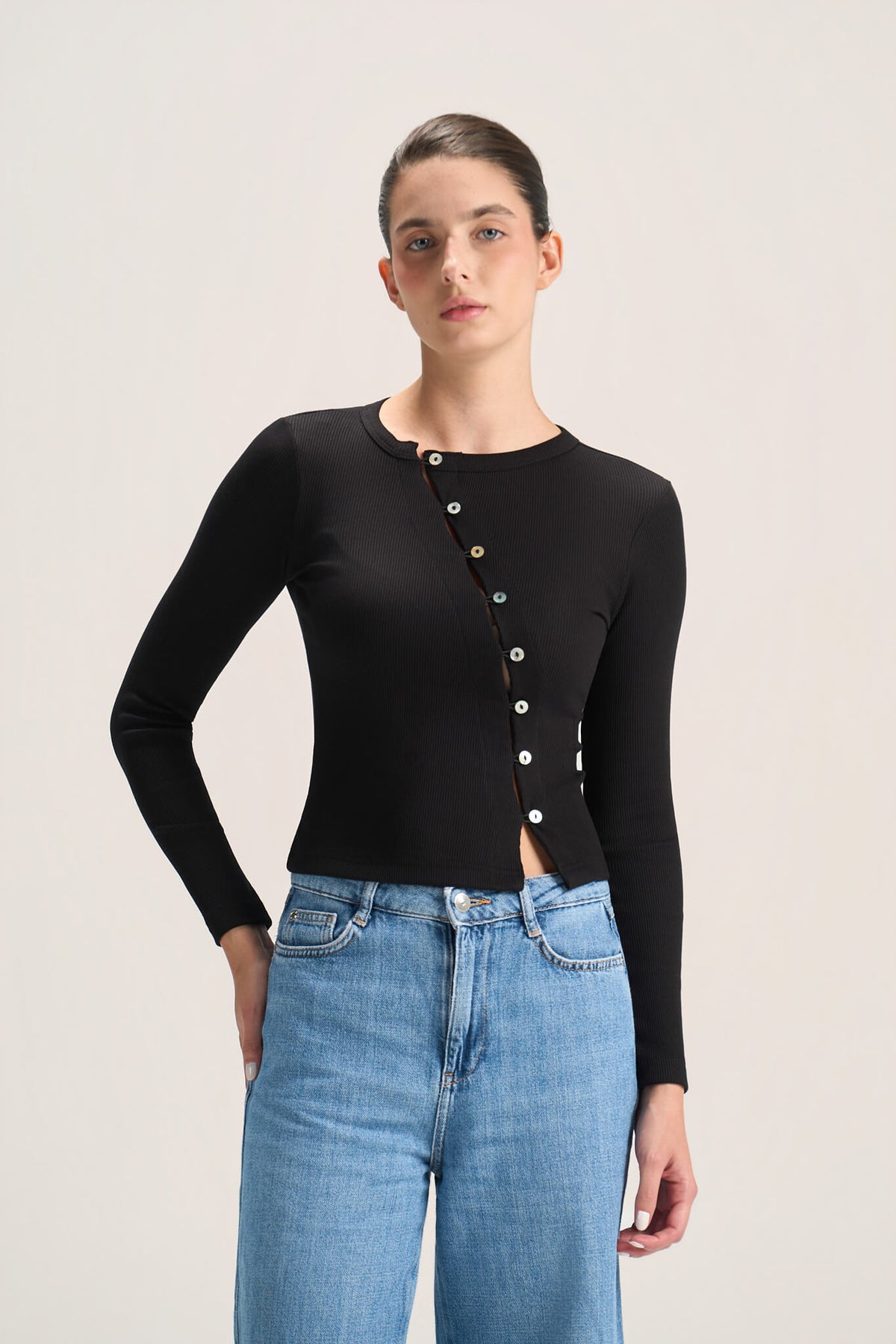 A model wearing a black long-sleeve knit top with a decorative button-down front and light blue jeans, photographed against a plain light gray studio background.