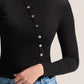 A close-up of a person wearing a black ribbed long-sleeve top with a vertical row of decorative buttons down the front, paired with blue jeans against a plain light background.