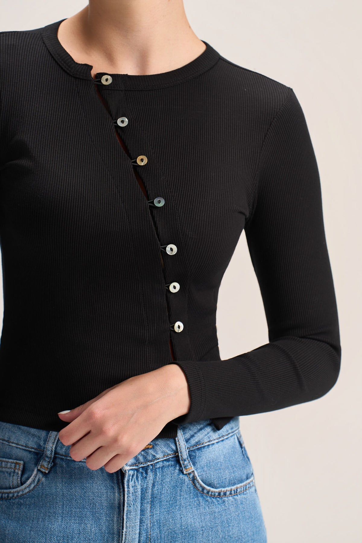 A close-up of a person wearing a black ribbed long-sleeve top with a vertical row of decorative buttons down the front, paired with blue jeans against a plain light background.