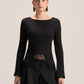 A model wearing a black long-sleeve knit top with a boat neckline, a sheer lace waist panel, and black draped trousers, photographed against a solid light beige background.