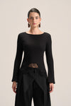 A model wearing a black long-sleeve knit top with a boat neckline, a sheer lace waist panel, and black draped trousers, photographed against a solid light beige background.
