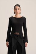 A model wearing a black long-sleeve knit top with a boat neckline, a sheer lace waist panel, and black draped trousers, photographed against a solid light beige background.