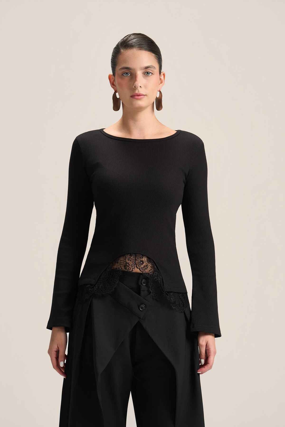 A model wearing a black long-sleeve knit top with a boat neckline, a sheer lace waist panel, and black draped trousers, photographed against a solid light beige background.