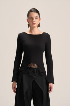A model wearing a black long-sleeve knit top with a boat neckline, a sheer lace waist panel, and black draped trousers, photographed against a solid light beige background.