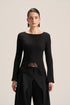 A model wearing a black long-sleeve knit top with a boat neckline, a sheer lace waist panel, and black draped trousers, photographed against a solid light beige background.