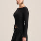 A woman modeling a black long-sleeve knit top with a boat neckline and a sheer lace side detail, paired with a black skirt, against a solid light beige background.