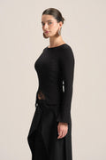 A woman modeling a black long-sleeve knit top with a boat neckline and a sheer lace side detail, paired with a black skirt, against a solid light beige background.