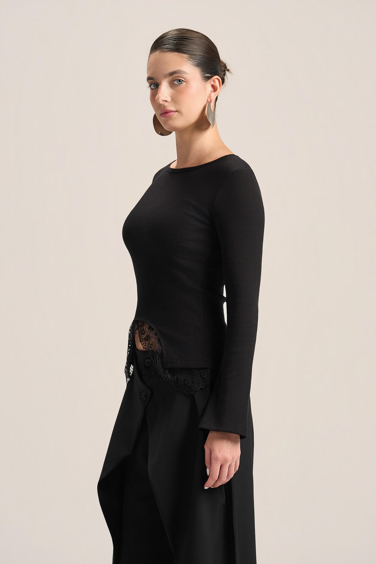 A woman modeling a black long-sleeve knit top with a boat neckline and a sheer lace side detail, paired with a black skirt, against a solid light beige background.