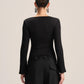 A model shown from the back wearing a black long-sleeve top with a boat neckline and black wide-leg trousers, set against a plain light beige studio background.