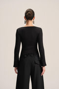 A model shown from the back wearing a black long-sleeve top with a boat neckline and black wide-leg trousers, set against a plain light beige studio background.