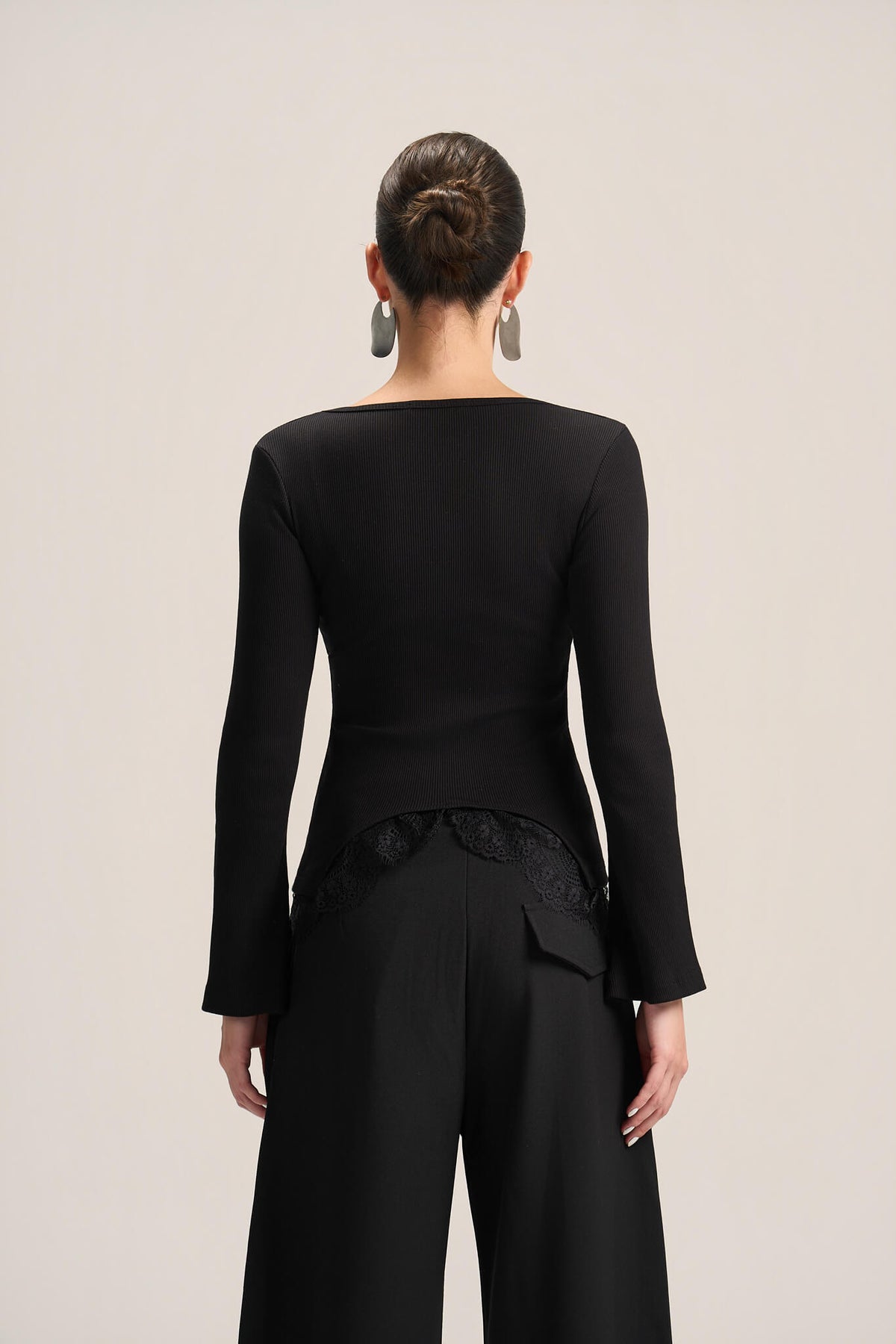 A model shown from the back wearing a black long-sleeve top with a boat neckline and black wide-leg trousers, set against a plain light beige studio background.