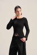 A model wearing a black long-sleeve boat-neck top with a unique asymmetrical hem and black wide-leg trousers, posed against a solid light beige background.