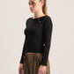 A woman with blonde hair in a ponytail is shown from the waist up, wearing a black long-sleeve crop top with decorative buttons on the shoulders and high-waisted brown trousers, set against a plain light gray studio background.