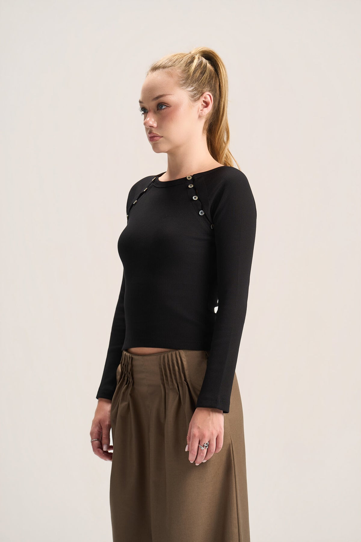 A woman with blonde hair in a ponytail is shown from the waist up, wearing a black long-sleeve crop top with decorative buttons on the shoulders and high-waisted brown trousers, set against a plain light gray studio background.