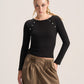 A woman modeling a fitted black long-sleeve top with decorative silver-tone studs along the shoulders, paired with brown pleated wide-leg pants, against a plain light gray studio background.