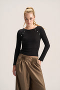 A woman modeling a fitted black long-sleeve top with decorative silver-tone studs along the shoulders, paired with brown pleated wide-leg pants, against a plain light gray studio background.