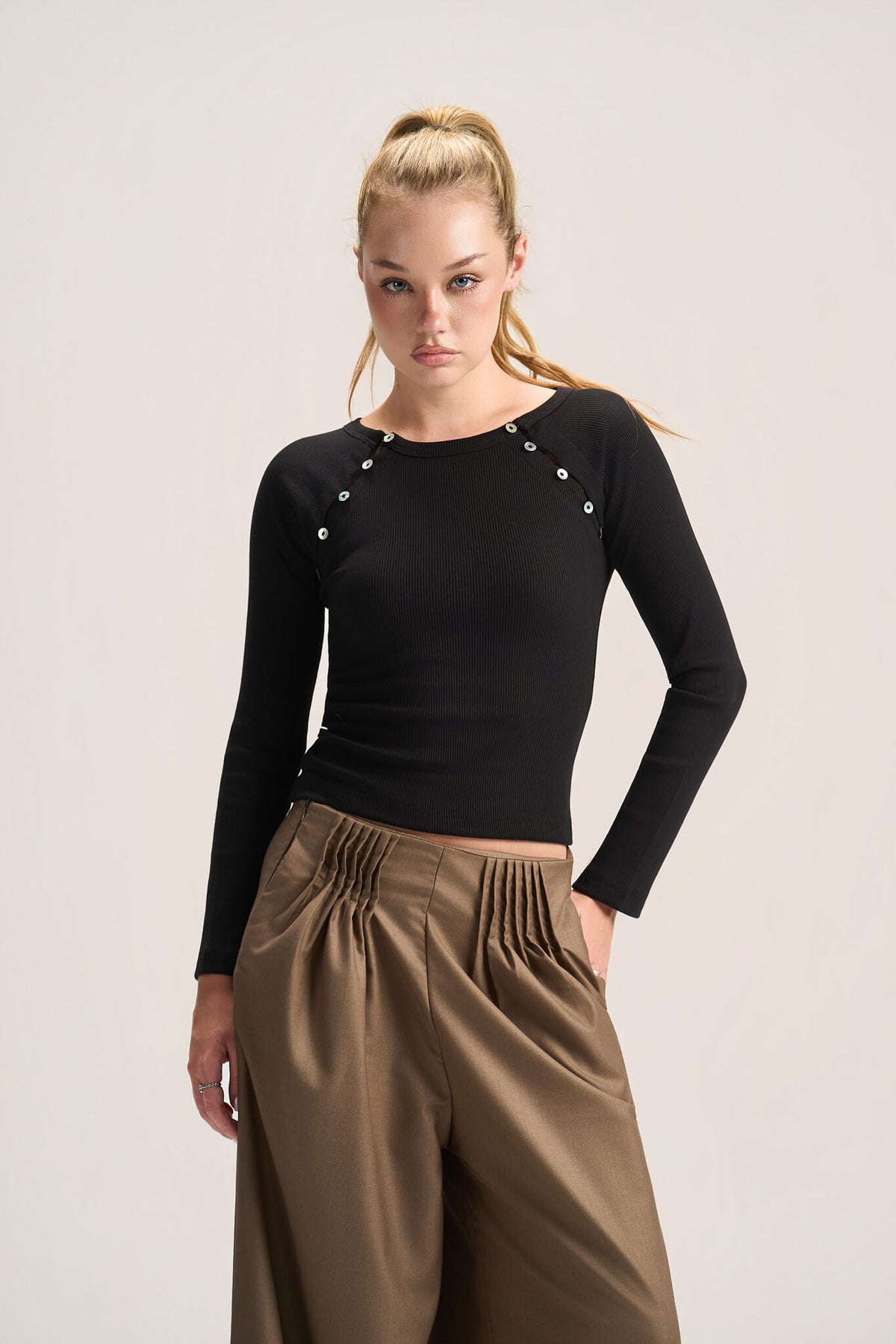 A woman modeling a fitted black long-sleeve top with decorative silver-tone studs along the shoulders, paired with brown pleated wide-leg pants, against a plain light gray studio background.