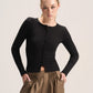 A blonde model wearing a fitted black long-sleeve button-front top and brown gathered wide-leg pants, standing against a plain light beige studio background.