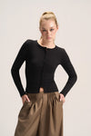 A blonde model wearing a fitted black long-sleeve button-front top and brown gathered wide-leg pants, standing against a plain light beige studio background.