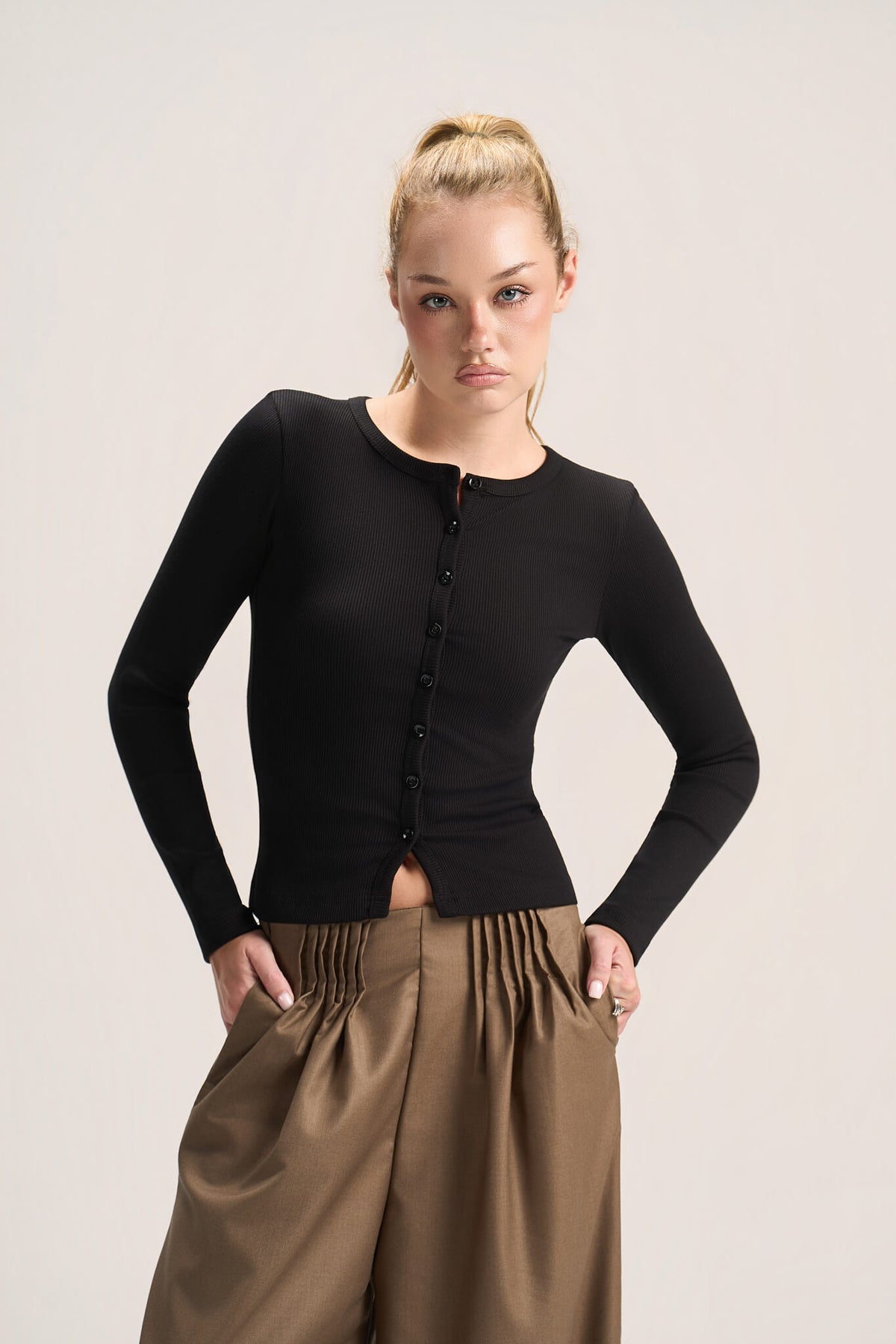 A blonde model wearing a fitted black long-sleeve button-front top and brown gathered wide-leg pants, standing against a plain light beige studio background.