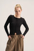 A blonde model wearing a fitted black long-sleeve button-front top and brown gathered wide-leg pants, standing against a plain light beige studio background.