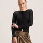A blonde model wearing a black long-sleeve button-front knit top and brown pleated wide-leg pants, standing against a light gray studio background.