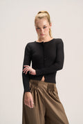 A blonde model wearing a black long-sleeve button-front knit top and brown pleated wide-leg pants, standing against a light gray studio background.