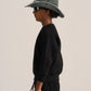 A child model shown in profile wearing a black sweatshirt and matching black pants, with a muted blue-green bucket hat and dark sunglasses against a plain light beige studio background.