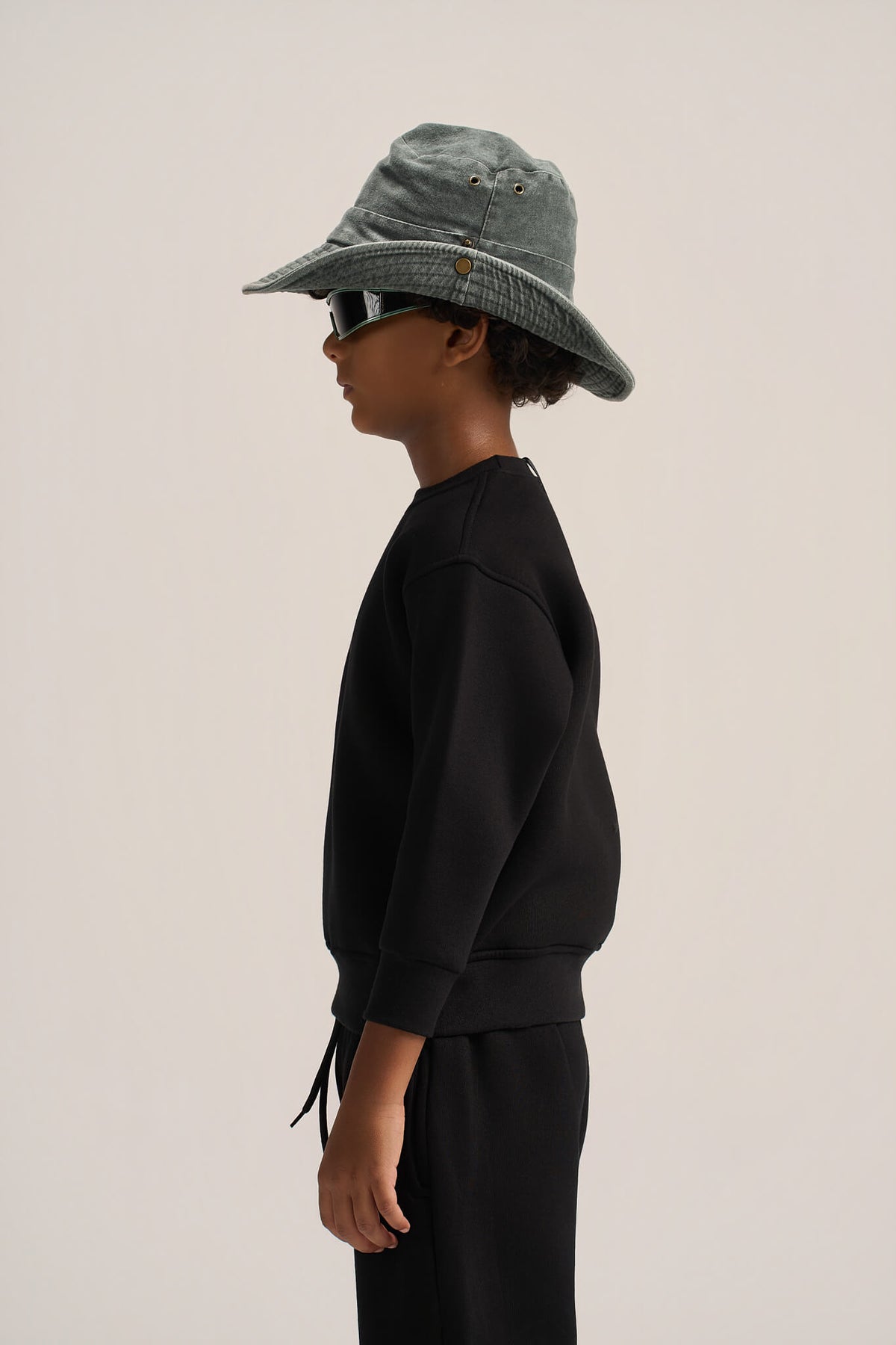 A child model shown in profile wearing a black sweatshirt and matching black pants, with a muted blue-green bucket hat and dark sunglasses against a plain light beige studio background.