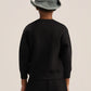 A child shown from the back wearing a black long-sleeve sweatshirt and black pants, with a gray-green bucket hat, against a plain light beige studio background.
