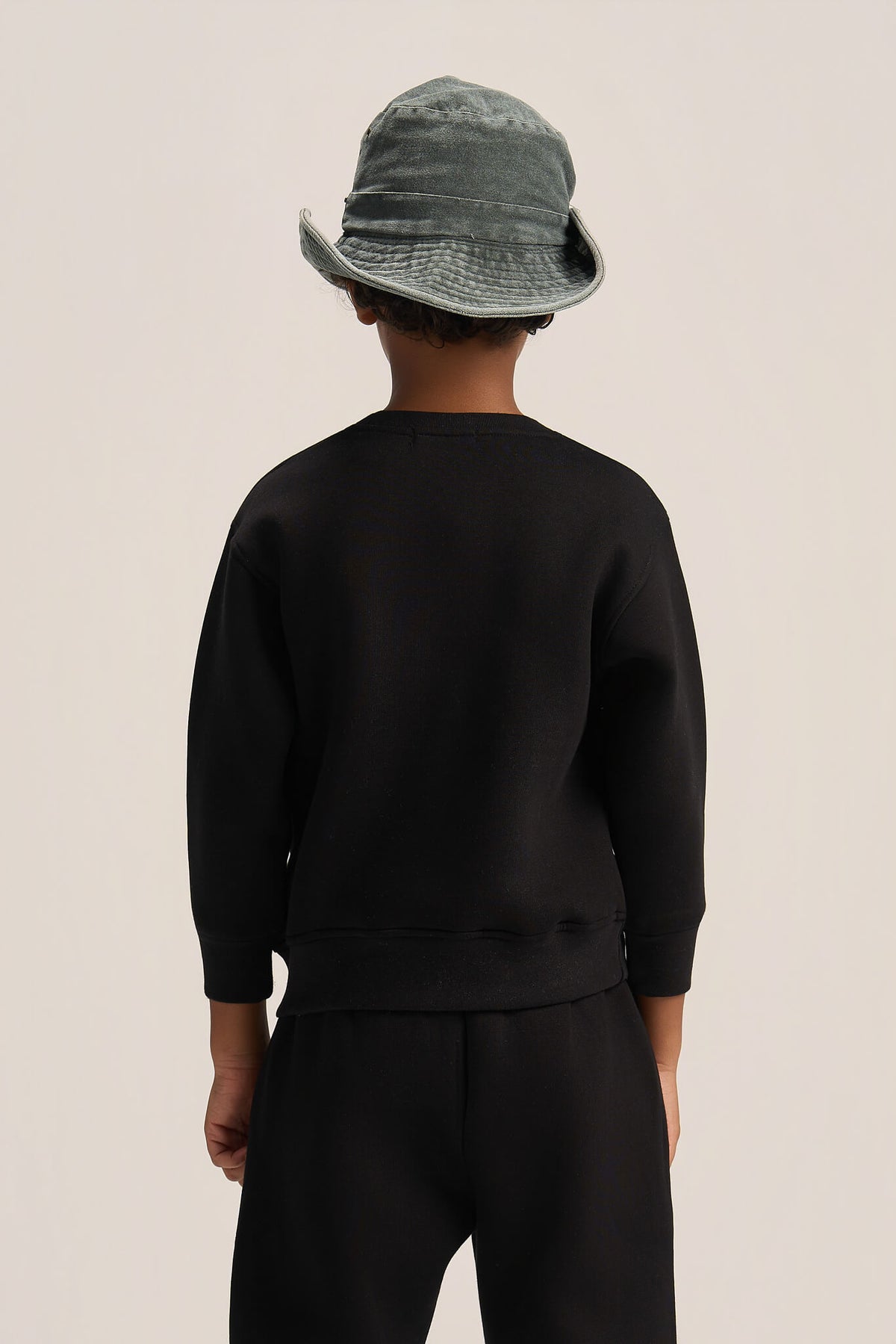 A child shown from the back wearing a black long-sleeve sweatshirt and black pants, with a gray-green bucket hat, against a plain light beige studio background.
